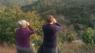 2 people standing on a hill and looking through binoculars at a lush green mountain range.