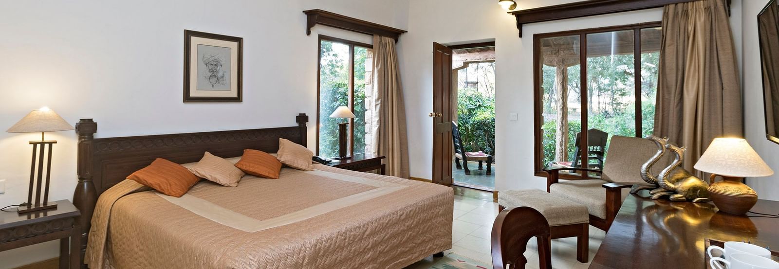 A bedroom with a double bed, side tables with lamps, desk with cups and decor, wooden chairs, patterned rug, ceiling fan, and doors leading outside - MANVÂR Shergarh The Desert Resort