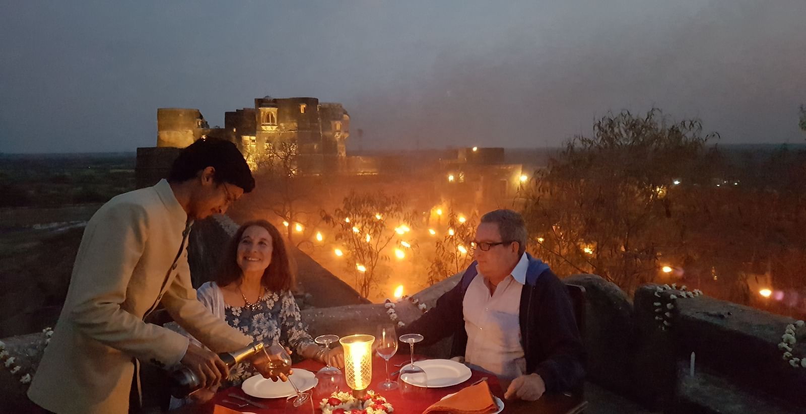 A waiter serves a meal to two people at a candlelit table on a rooftop at dusk, with lights visible in the distance- Shahpura Bagh.