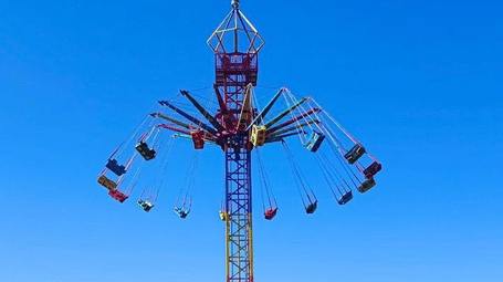 Outdoor vertical spinning ride with seats on arms extending from a tall central pole against clear blue sky.