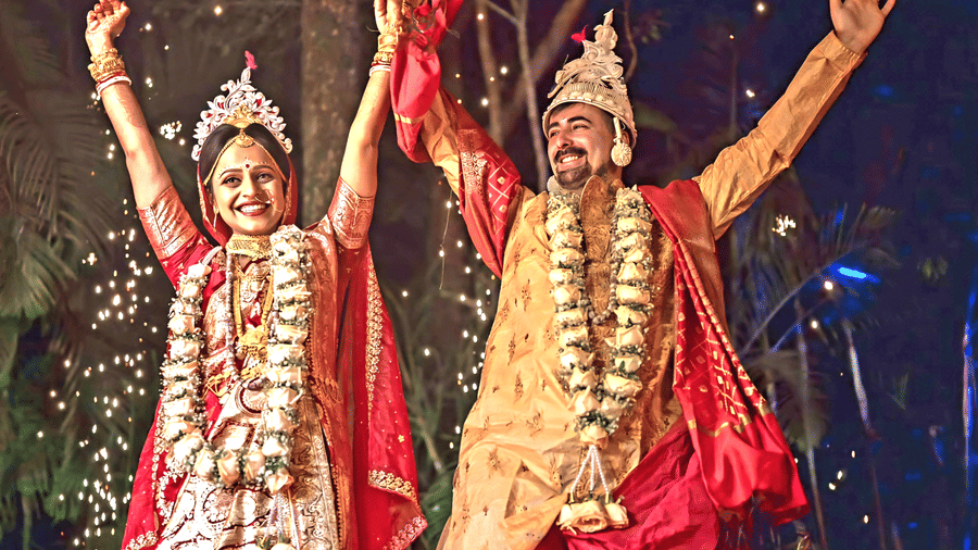 A bride and groom in traditional wedding attire celebrating joyfully at Vedic Village Spa Resort with hands raised under colorful stage lighting and festive decorations in the background.