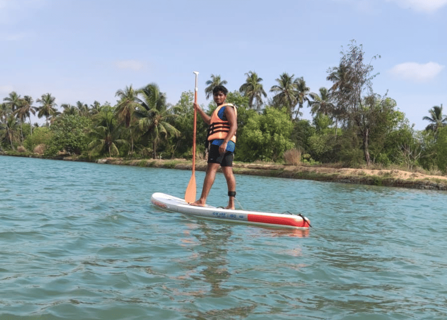 One person standing on paddle-boards in the river under a bright blue sky, holding paddles at Paradise Lagoon Resort, Udupi.