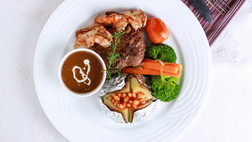 A plated dish features meat, vegetables, and a sauce drizzle on a white plate, presented with a patterned cloth.