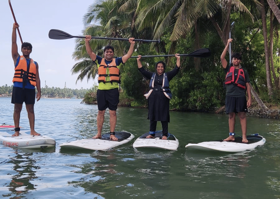 Three people standing on paddle-boards in the river near palm trees, balancing with paddles in hand at Paradise Lagoon Resort, Udupi.