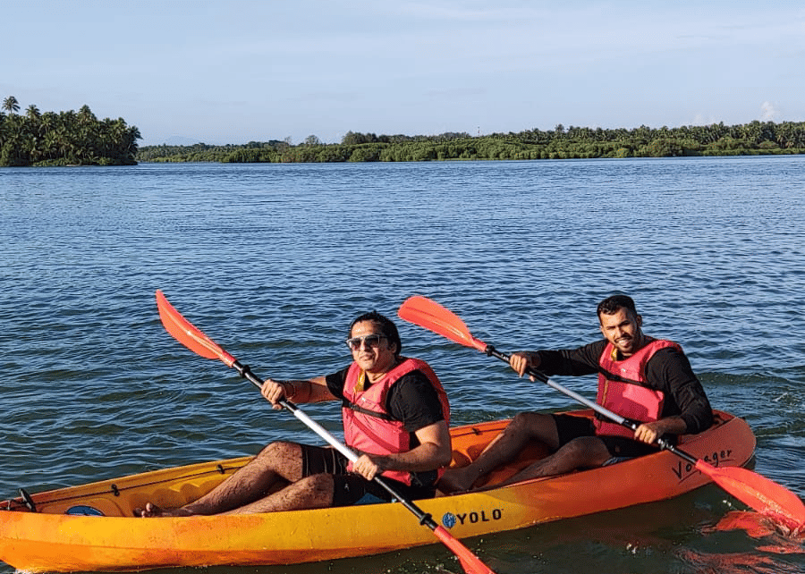 Two people in orange life jackets paddling an orange kayak on a calm river under a clear blue sky at Paradise Lagoon Resort, Udupi.