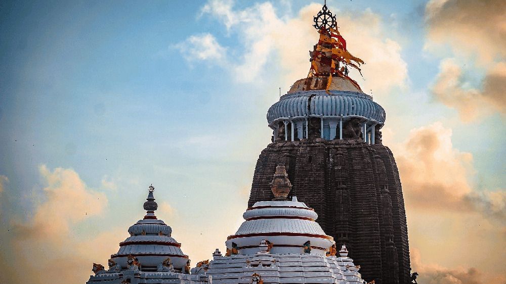  A low-angle photo of the tall, dark-stone Jagannath Temple tower against a cloudy sky at sunset, with lower white structures in the foreground at Swosti Premium Beach Resorts, Puri.