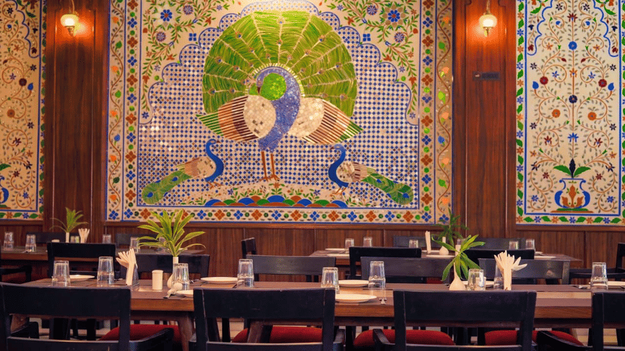 A restaurant dining table with dark chairs, in front of a wall featuring a large mosaic panel depicting a peacock at Amantra Shilpi Resort, Udaipur.
