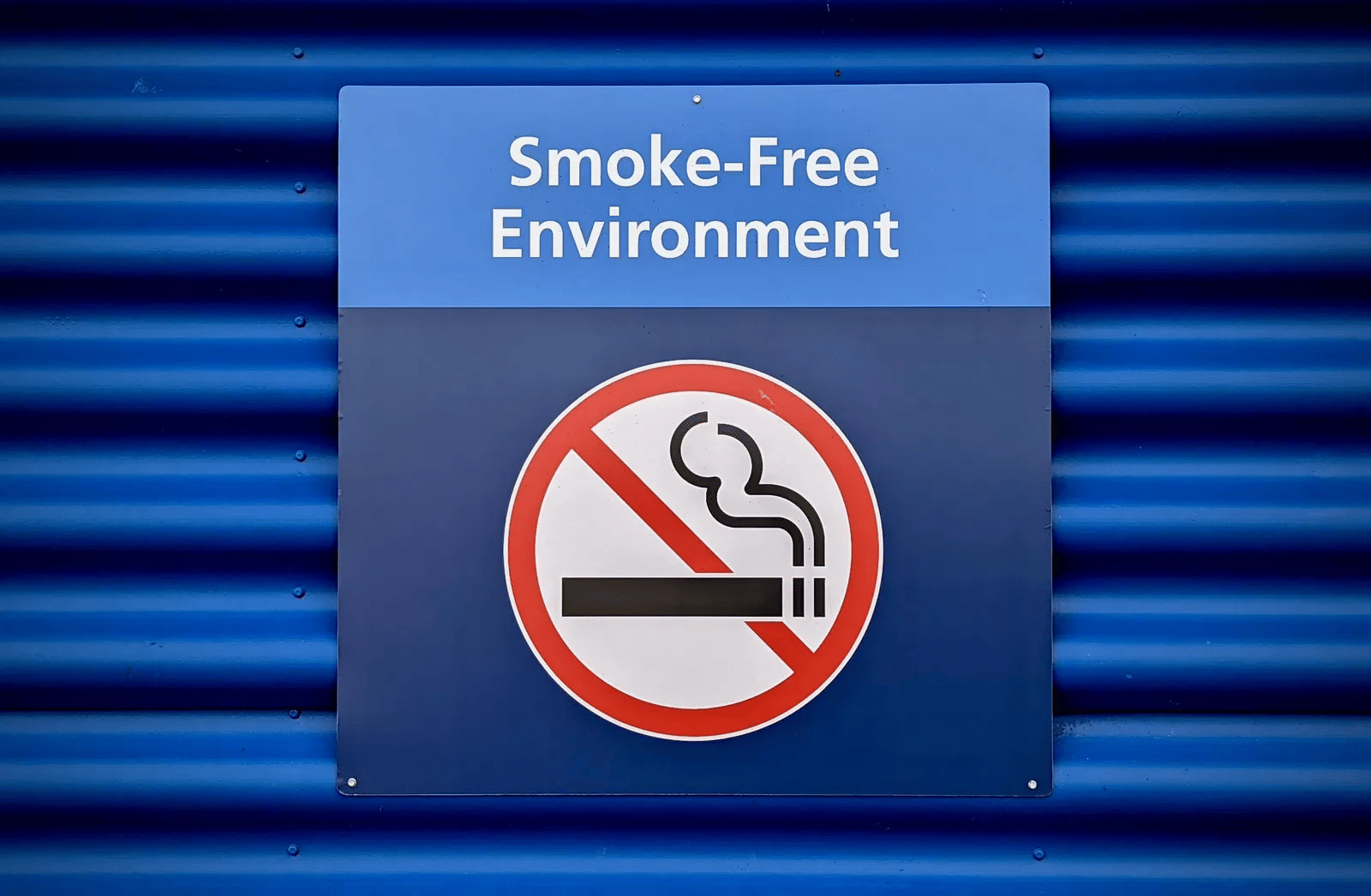 A square blue sign featuring a no-smoking symbol and the text 'Smoke-Free Environment' mounted on a blue corrugated wall.