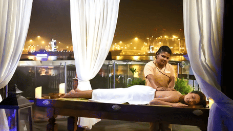 Nighttime spa treatment on an outdoor deck with white sheer curtains, where a masseuse massages a client wrapped in a white towel.