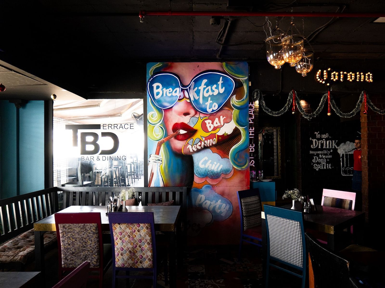 A colourful indoor dining area with a large pop art mural of a woman wearing sunglasses and drinking through a straw. The mural includes the words Breakfast to Bar. The restaurant is called TBD Bar & Dining. 