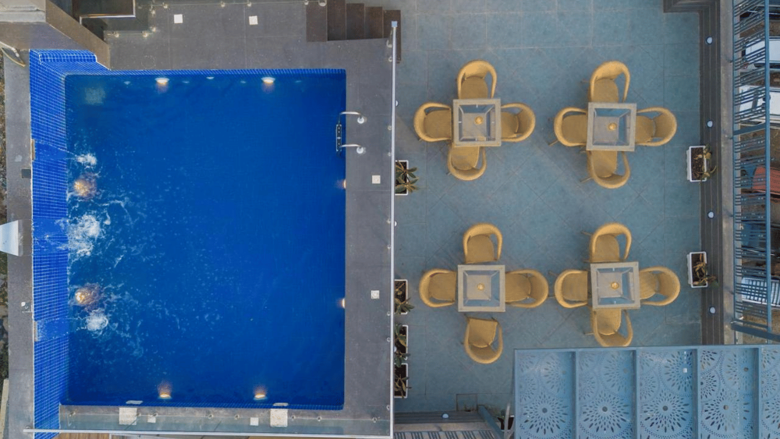 An aerial view of a rooftop at Perfectstayz Premium Laxmi Heritage with a rectangular swimming pool, and 4 groups of wicker chairs and tables.