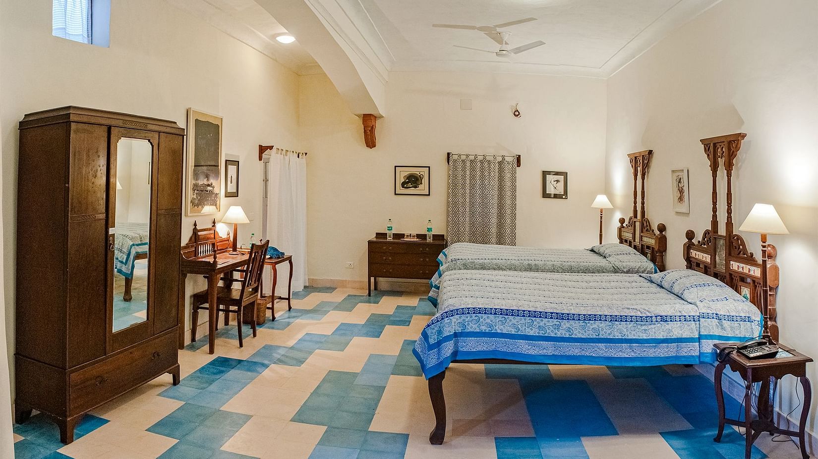 A room with two single beds, a wooden wardrobe, a desk with chairs, and a patterned floor in Tijara Fort-Palace - 19th Century, Alwar.
