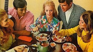 A group of people, including adults and children, are gathered around a table enjoying a fondue meal, with a fondue pot and various dipping items visible.