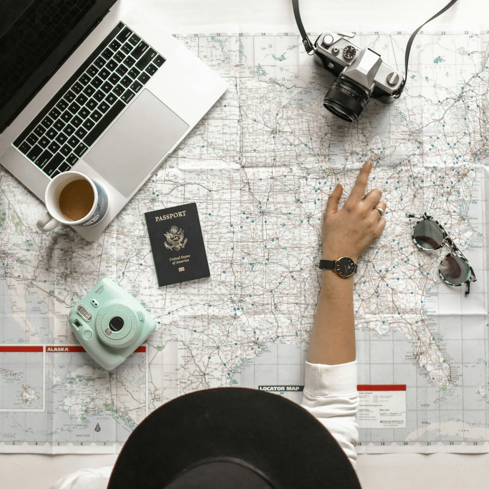 Overhead view of a person wearing a hat and pointing at a map with a laptop, camera, passport, sunglasses, coffee cup, and a vintage camera placed on the table.