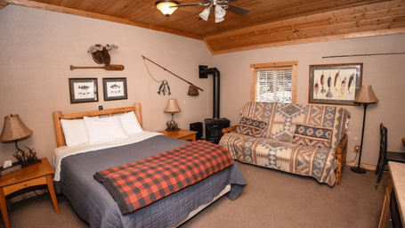 a Room with a bed, plaid blanket, patterned couch, wood ceiling, fishing decor, and two framed pictures at Shaver Lake Village Hotel.