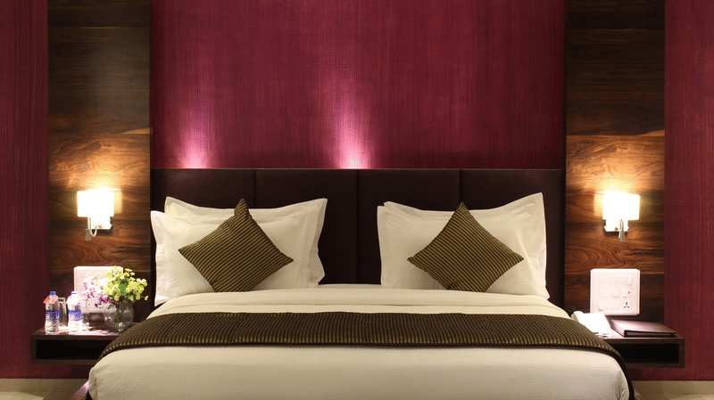 A room showcases a king size bed with a dark purple headboard, bedside tables with lamps, and elegant bedding.