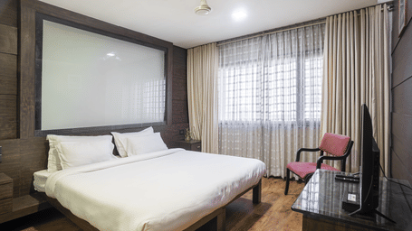 A spacious room with a king size bed, beige curtains, dark wall panelling, a pink armchair, and a T.V. at Infiniti Hotel, Indore.