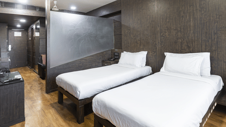 A room with two single beds, dark textured walls, a ceiling light, and a compact open wardrobe unit at Infiniti Hotel, Indore.