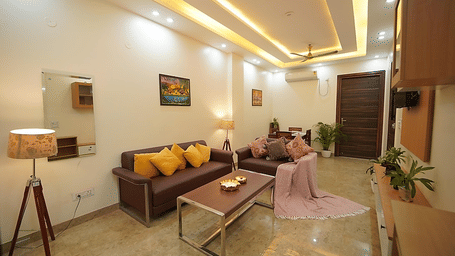 Modern serviced apartment living room at Lime Tree Hotels & Service Apartment Private Limited with plush sofas, warm ceiling lighting, marble flooring, wooden furnishings, indoor plants, and cosy decor.