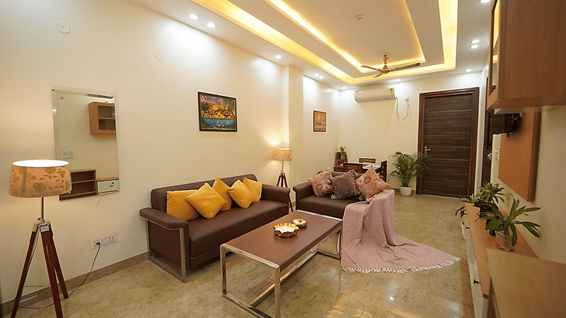 Living room of Lime Tree Hotels & Service Apartment Private Limited featuring warm cove lighting, a sofa with yellow cushions, and a floor lamp.