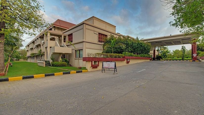 Exterior view of The Ummed Ahmedabad featuring a spacious driveway, lush greenery, and a clear blue sky.