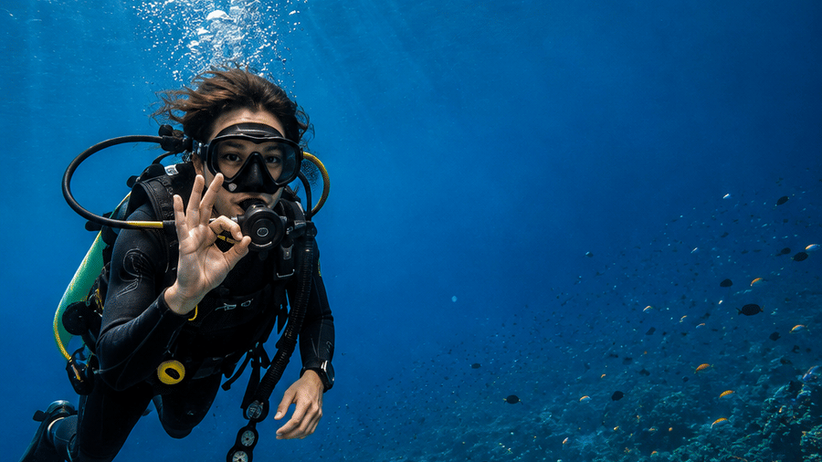 A scuba diver swimming underwater above a coral reef in clear blue ocean water.