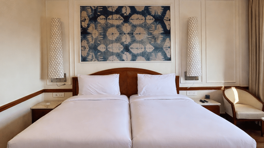 Twin beds with white linens and a central decorative headboard artwork sit beneath a ceiling fan in the Deluxe Twin room at Clarks Amer, Jaipur.
