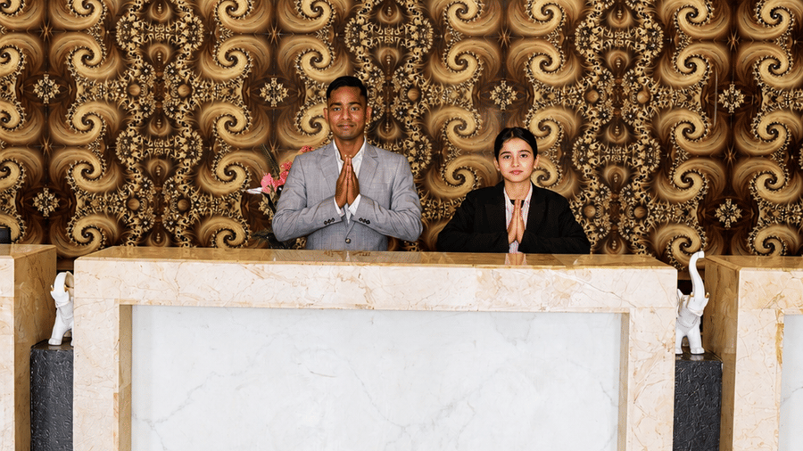 Front desk at Golden Tulip Kukas Jaipur with two staff members at a white reception counter, decorative dark patterned wall backdrop, and marble flooring.