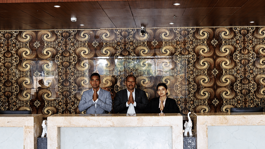 Reception area at Golden Tulip Kukas Jaipur featuring a white front desk, ornate dark patterned wall, marble floors, and warm overhead lighting.