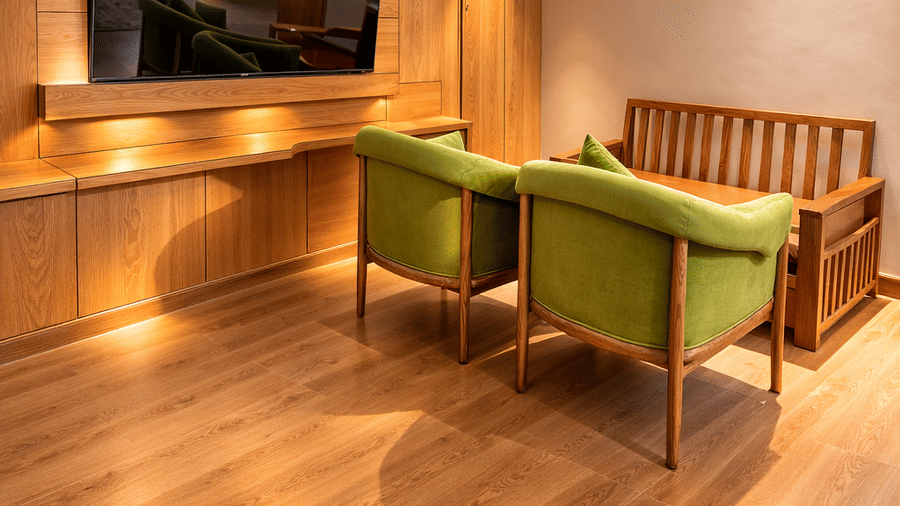 A vertical view of a cozy seating area within a suite at Mountnoor Sarovar Premiere, showcasing wood flooring, two armchairs, a wall-mounted television, and warm ambient cove lighting along the ceiling.
