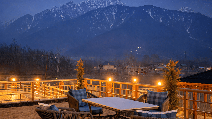 Facade of Mountnoor Sarovar Premiere resort in Pahalgam, a dining table and chairs sit on a stone patio, overlooking a breathtaking view of a star-filled sky over the dark silhouettes of the mountain peaks.