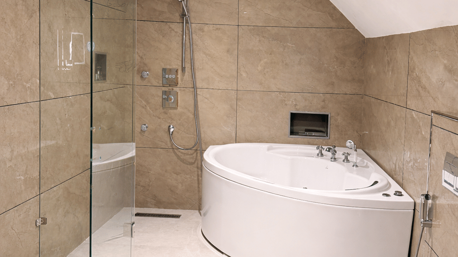 An ensuite bathroom at Mountnoor Sarovar Premiere showing a glass shower partition next to a curved white bathtub and a toilet.