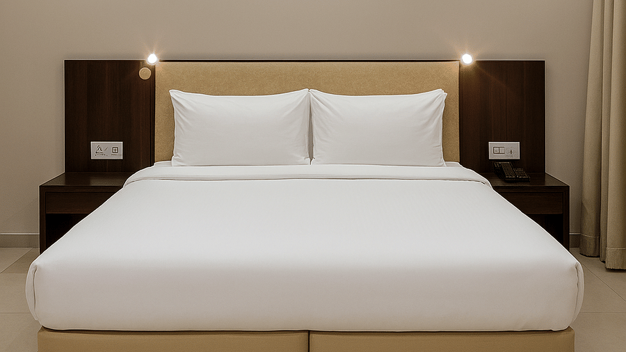 A centred view of a double bed at the Superior Room in Tropicana Resort & Spa, Alibaug, featuring bedding, a sleek dark wood headboard, and built-in bedside reading lights.
