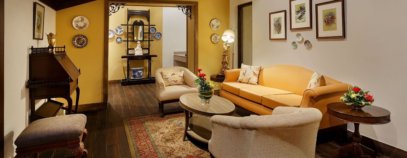 An image of the living area with Sofas that could seat 5 people, spaciously arranged furniture, multiple wall paintings, and a slanted wooden ceiling which accentuates the ambience.