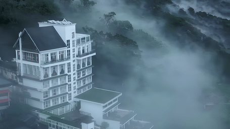 A tall, modern white and dark-roofed hotel building on a steep hillside, partially obscured by dense fog and mist.