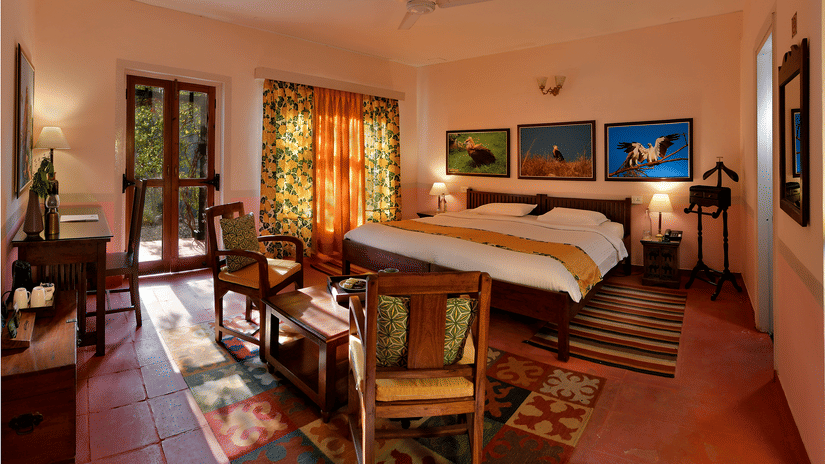 Bright heritage bedroom with wooden bed seating area patterned floor rugs artwork and long windows allowing natural light throughout the room at Neemrana’s Glasshouse on The Ganges.