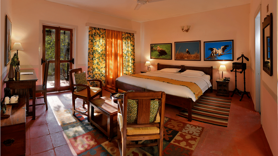 Bright heritage bedroom with wooden bed seating area patterned floor rugs artwork and long windows allowing natural light throughout the room at Neemrana’s Glasshouse on The Ganges offering a welcoming and restful environment