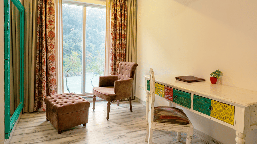 A room featuring an upholstered armchair, an ottoman, a painted wooden desk and a chair, a framed mirror on a wall, and a big window with a view of the river and trees outside | Summit by the Ganges Beach Resort & Spa, Rishikesh