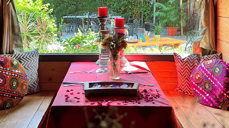An indoor dining nook with a table set with crockery, floral arrangements, candles, and decorative cushions, overlooking a garden.