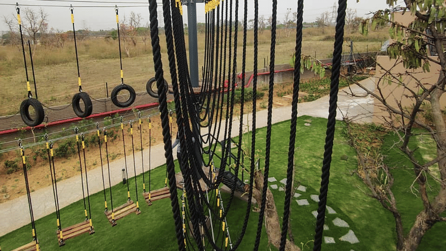An elevated view featuring a narrow rope bridge with vertical safety ropes suspended over a green lawn at Hotel Sonar Bangla, Ajodhya Hills.
