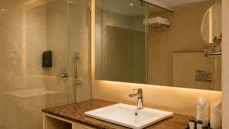 A bathroom vanity with a sink, a wide mirror, under-counter shelves with towels, and a glass shower cubicle at Pride Premier Greater Noida.