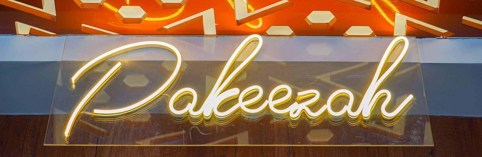 A glowing neon sign displaying Pakeezah in a cursive script is mounted on a patterned wall.