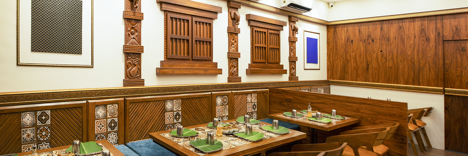 An image of a restaurant with wooden furnishings, table has three chairs on one side and a sofa on the other side - Kamats India