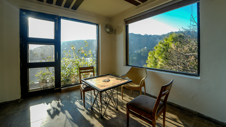 An image of the indoor game zone featuring a carrom board placed on a table with two chairs on either side and an a glass door and window overlooking the view - DLS Whispering Winds