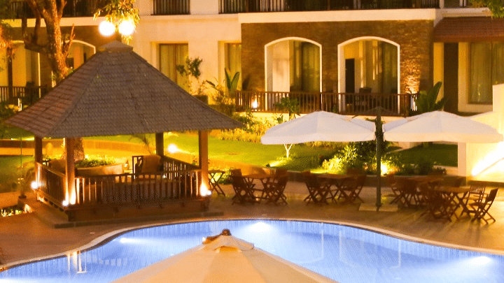 Exterior view of a resort with a large swimming pool, a thatched gazebo, dining tables with umbrellas, and a three-story hotel building in the background at dusk.
