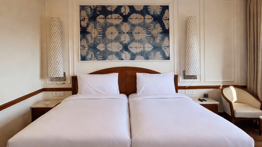 Twin beds with white linens and a central decorative headboard artwork sit beneath a ceiling fan in the Deluxe Twin room at Clarks Amer, Jaipur.