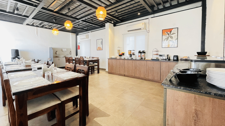 Dining area at Hotel Kyzen Smart Stays Madhapur featuring wooden tables, chairs, buffet counter along the wall, pendant lights, and large windows with natural light.