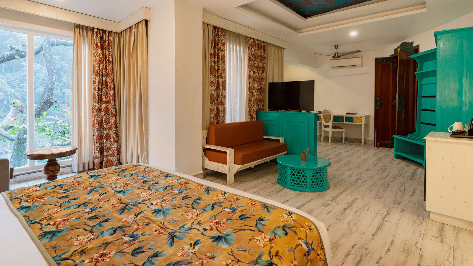 A spacious suite featuring a bed with a floral runner, a multi seater sofa, a wardrobe, a television unit, and a writing desk with a chair | Summit by the Ganges Beach Resort & Spa, Rishikesh