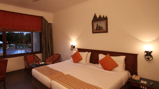 The dusk view of Deluxe Twin Pool View at Clarks Khajuraho, showcasing a large bed and accented pillows featuring dark wood furniture, a ceiling fan, and 2 wall-mounted sconces providing a warm glow. 