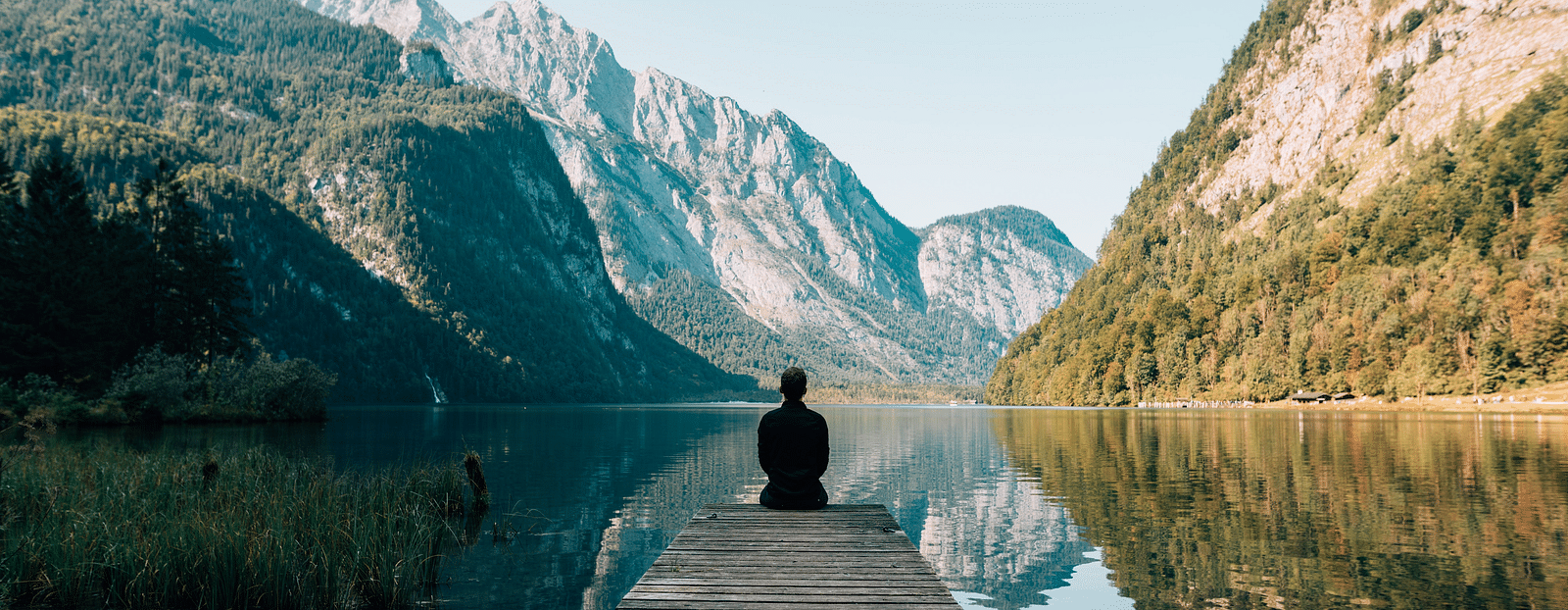 Person seated on a bench, admiring mountains reflected in the serene river.